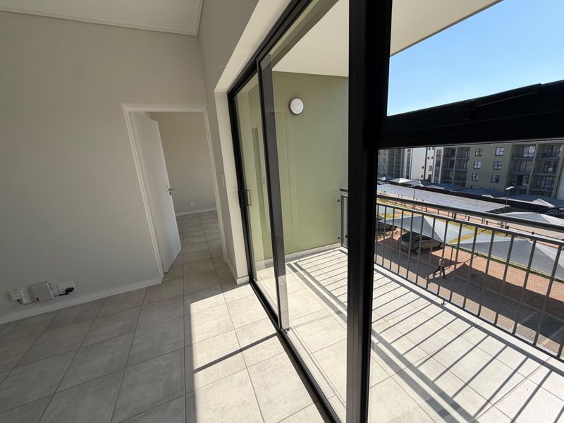 A bright, empty balcony with tiled flooring and metal balustrades, accessible via large sliding glass doors from the interior living space.