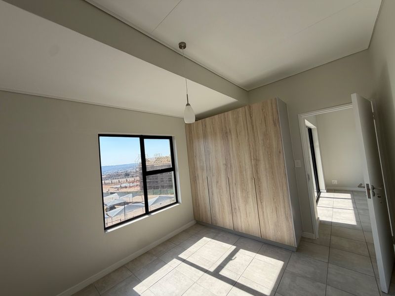 A bright, modern bedroom featuring neutral tiled flooring, built-in wood-finish cupboards, and an aluminium-framed window with natural light.