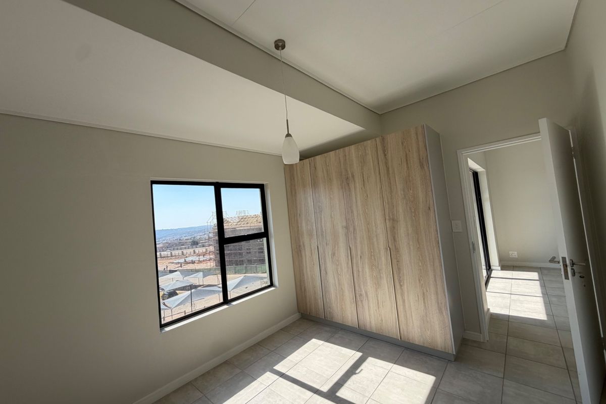 A bright, modern bedroom featuring neutral tiled flooring, built-in wood-finish cupboards, and an aluminium-framed window with natural light.