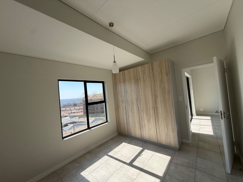 A bright, modern bedroom featuring neutral tiled flooring, built-in wood-finish cupboards, and a large window with natural light.