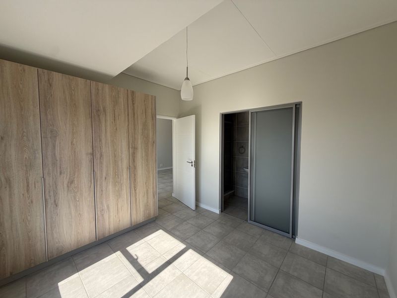 A bright, empty bedroom featuring modern wood-finish built-in cupboards, neutral tiled flooring, and direct access to an en-suite bathroom.