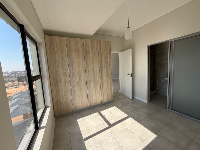 A bright, modern bedroom featuring built-in wood-finish cupboards, neutral tiled flooring, and an en-suite bathroom, offering a clean and functional living space.