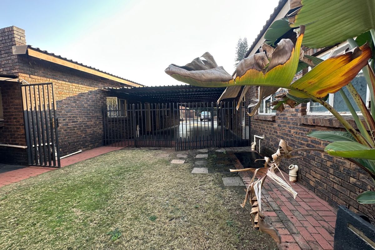 Rear exterior view of the property featuring a brick facade, a covered carport area, a metal security gate, and a small lawn with paved pathways.