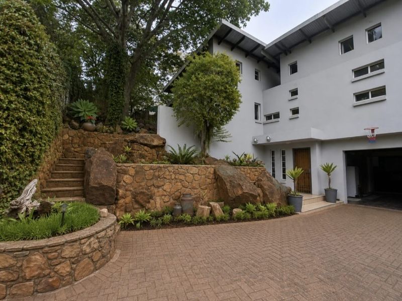 Front exterior view featuring a paved driveway, multi-level stone retaining walls, landscaped garden, and a multi-story house with a garage.
