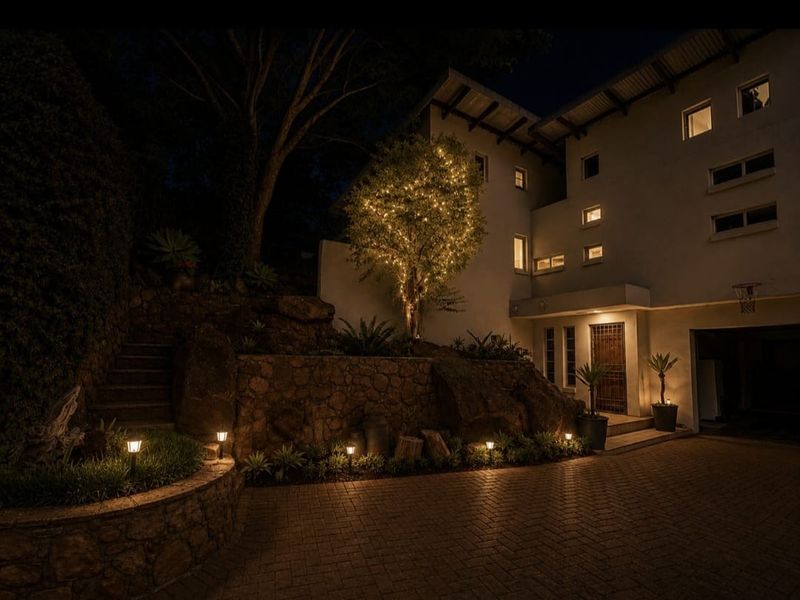 A well-lit front exterior view of the property featuring a stone retaining wall, paved driveway, and landscaped garden area at night.