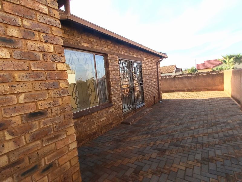 A rear exterior view featuring a paved patio area, face brick facade, and secure metal gates leading into the home.