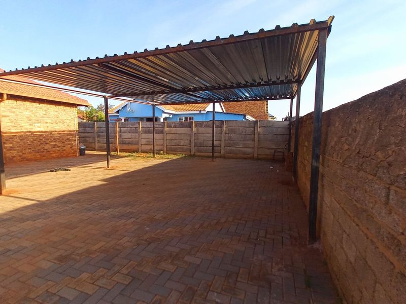 An open-sided carport structure featuring a corrugated metal roof supported by steel posts, set over a paved area with perimeter boundary walls.