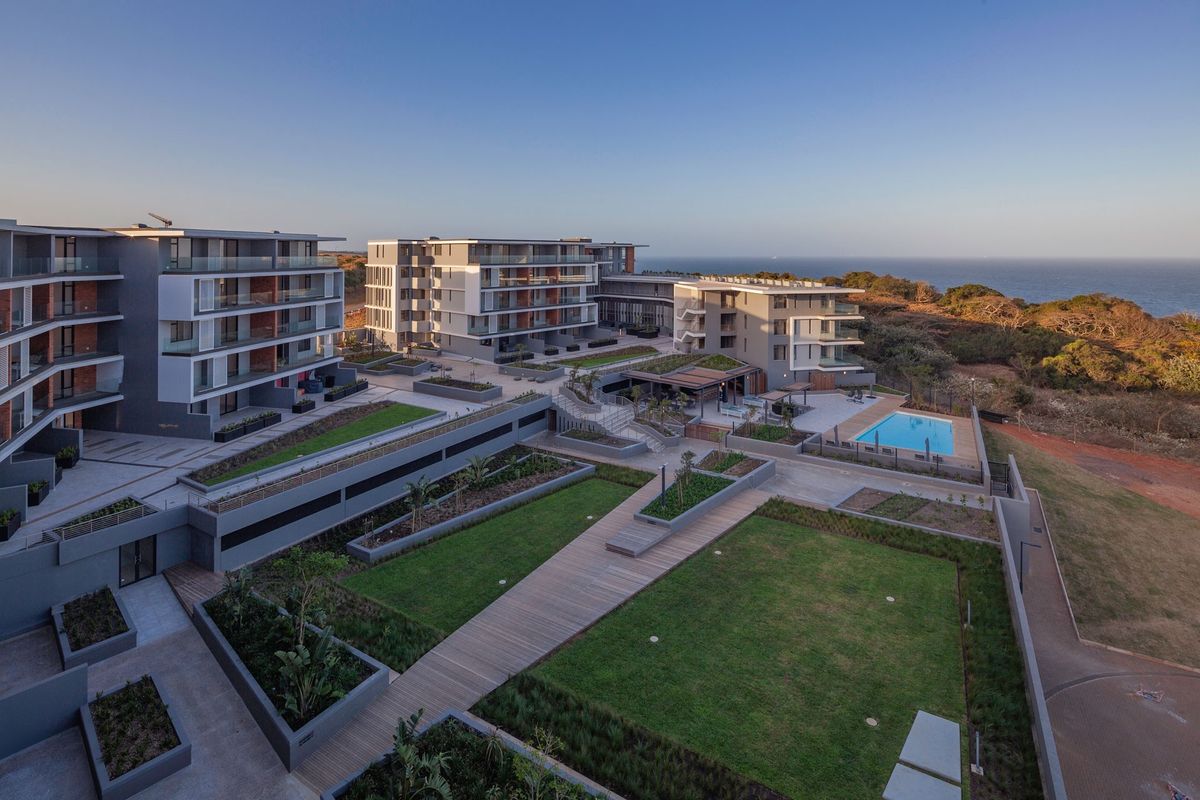 Aerial view of a modern apartment complex featuring landscaped gardens, paved walkways, a swimming pool, and ocean views.