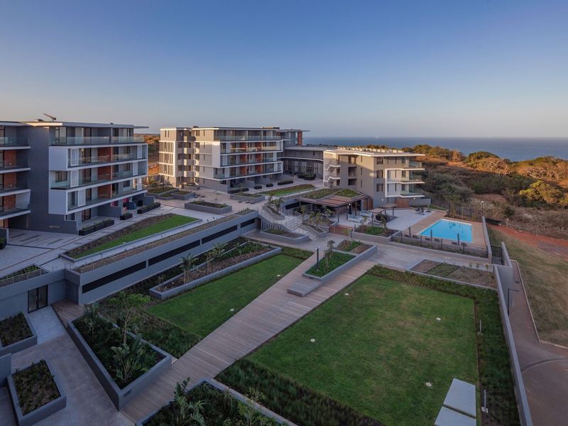 Aerial view of a modern apartment complex featuring landscaped gardens, paved walkways, a swimming pool, and ocean views.