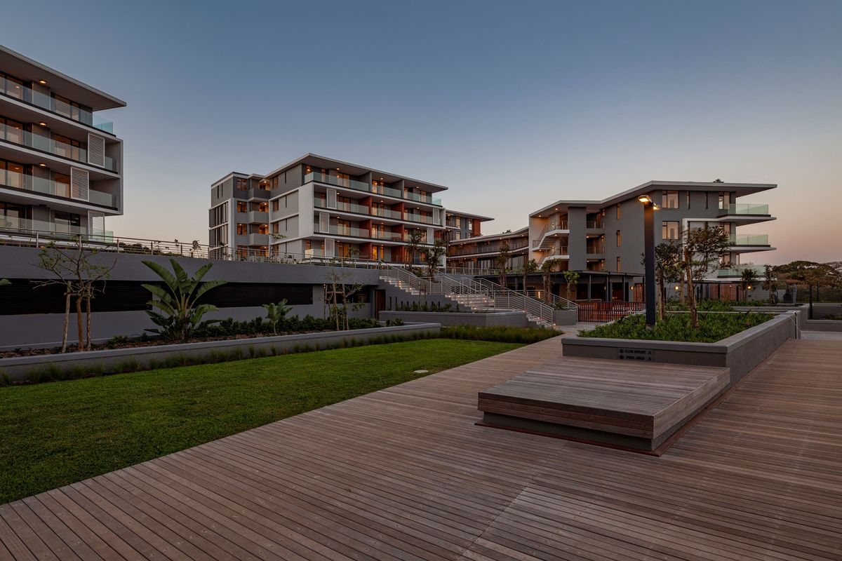 A modern apartment complex exterior featuring extensive timber decking, landscaped garden areas, and contemporary multi-story residential buildings with glass-fronted balconies.