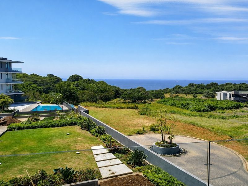 Elevated view overlooking a landscaped garden, swimming pool, and paved driveway with a scenic ocean horizon in the background.
