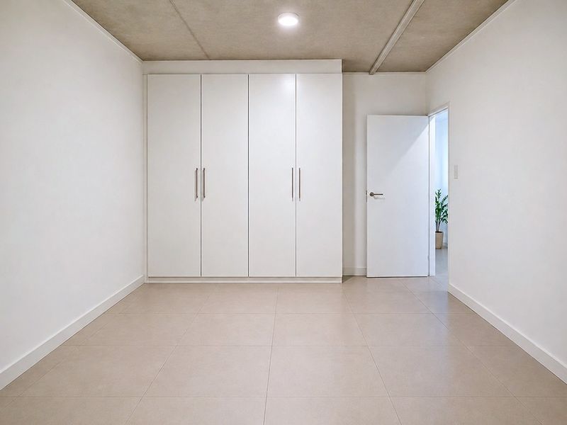 A clean, minimalist bedroom featuring neutral tiled flooring, a modern built-in wardrobe, and an exposed concrete ceiling.