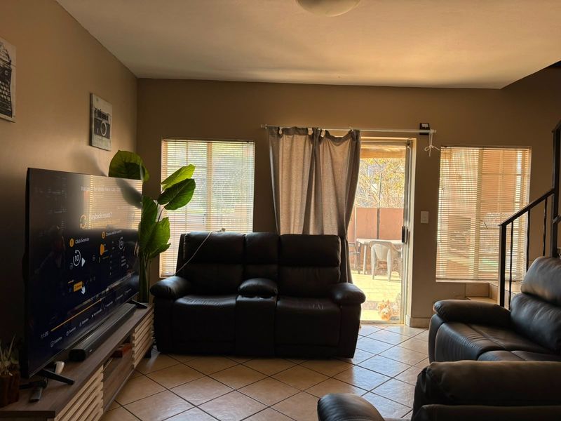 A living room featuring tiled flooring, large windows with venetian blinds, and a sliding glass door providing access to the outdoor patio area.