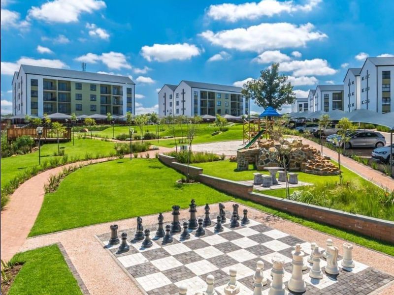 Well-maintained communal garden area featuring a large outdoor chess board, landscaped lawns, paved walkways, and a children's play area within a residential complex.