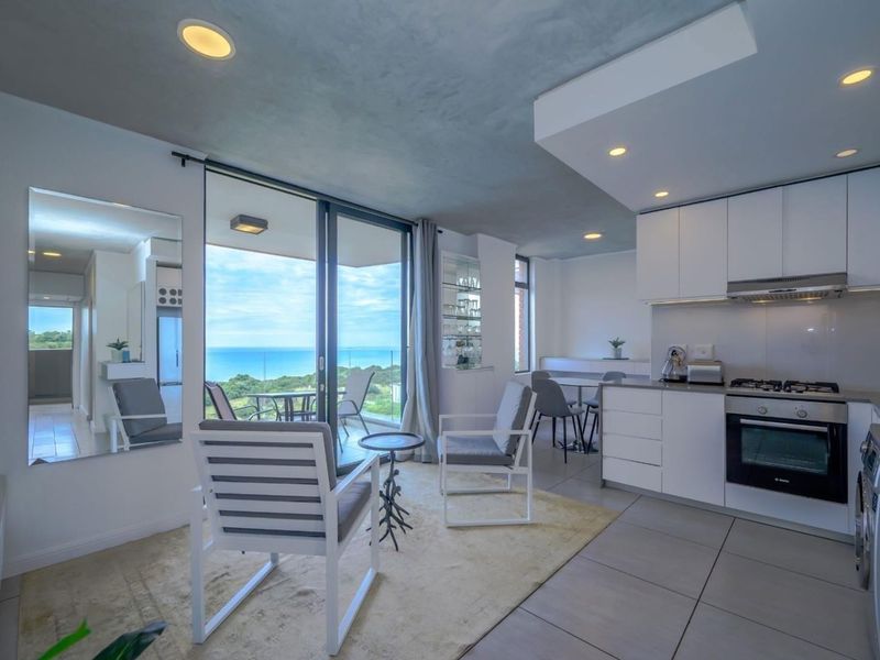 Modern open-plan kitchen and living area featuring sleek white cabinetry, integrated appliances, and large sliding doors opening to a scenic coastal view.