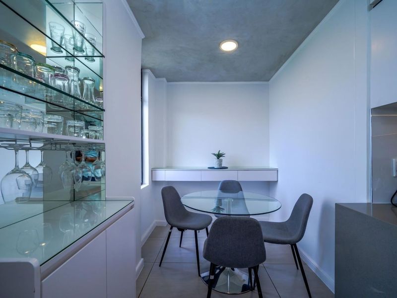 A modern dining area featuring a built-in glass display cabinet, floating shelf, recessed ceiling lighting, and neutral tiled flooring.