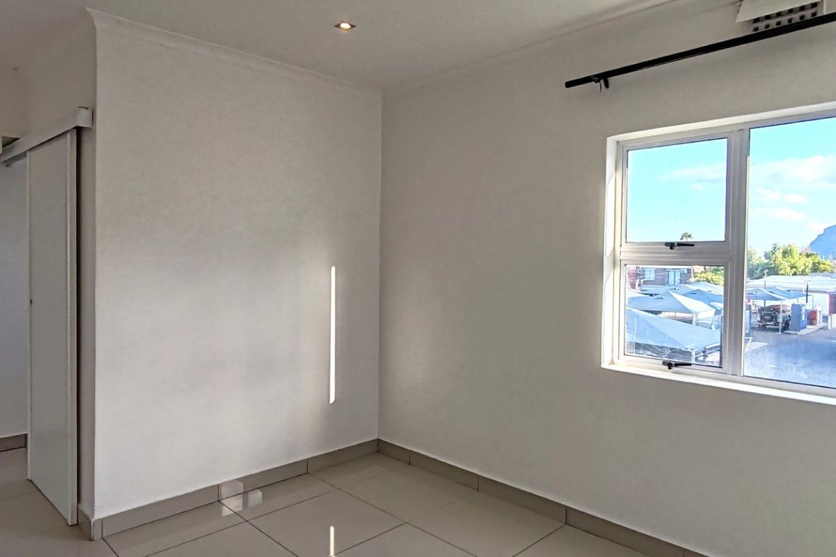A bright, empty bedroom featuring neutral tiled flooring, recessed downlighting, and a large window with views of the surrounding area.