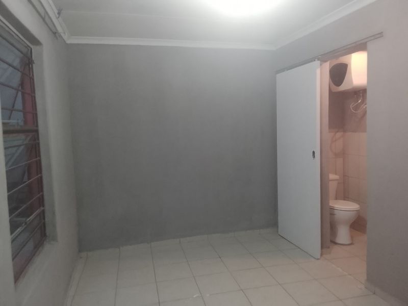 An empty bedroom featuring tiled flooring, a window with security bars, and an attached en-suite bathroom with a toilet and geyser.