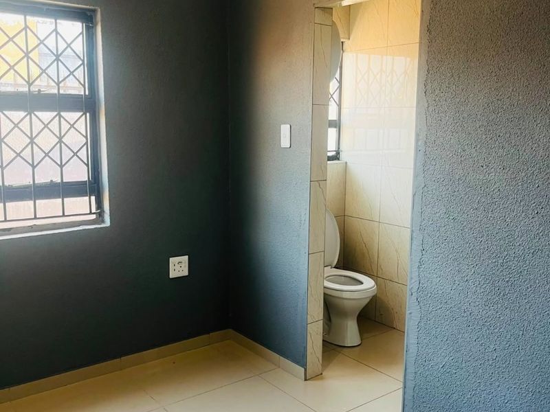 An empty bedroom featuring tiled flooring, security bars on the window, and an en-suite toilet area with tiled walls.
