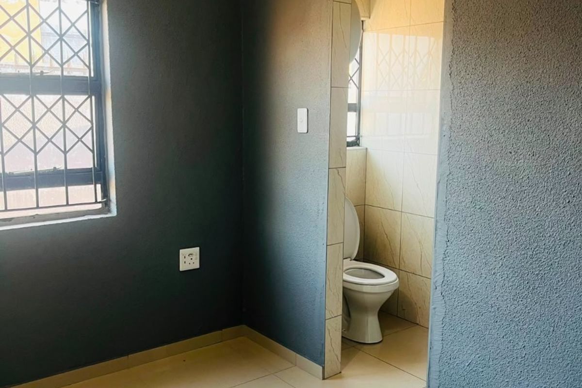 An empty bedroom featuring tiled flooring, security bars on the window, and an en-suite toilet area with tiled walls.