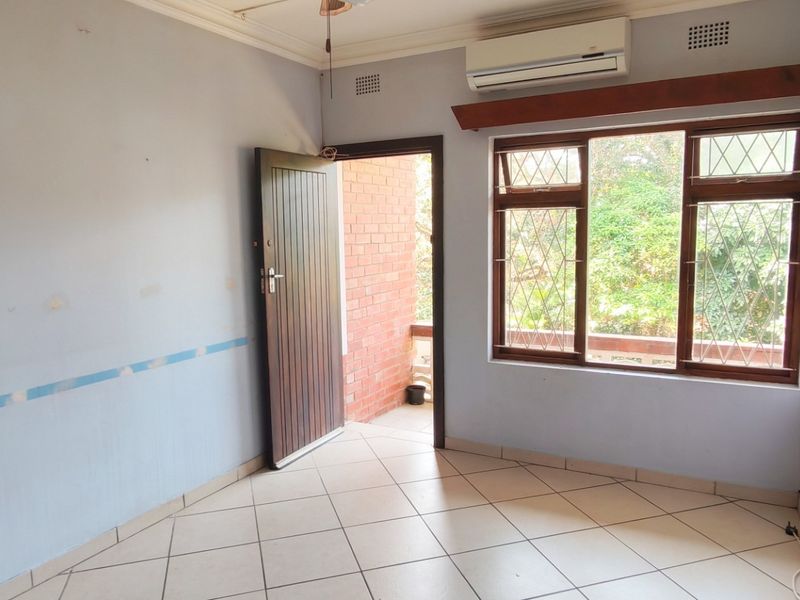 An empty bedroom featuring tiled flooring, an air conditioning unit, and wooden window frames with security bars, leading out to an exterior area.
