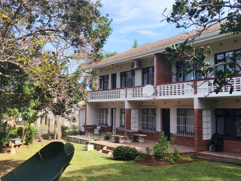 Rear view of a two-story apartment building featuring a ground-level patio, upper-level balcony, brick facade, and established garden surroundings.