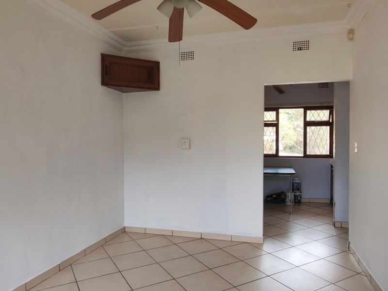 An empty living area featuring tiled flooring, a ceiling fan, and a corner wall cabinet, leading through to an adjacent room.