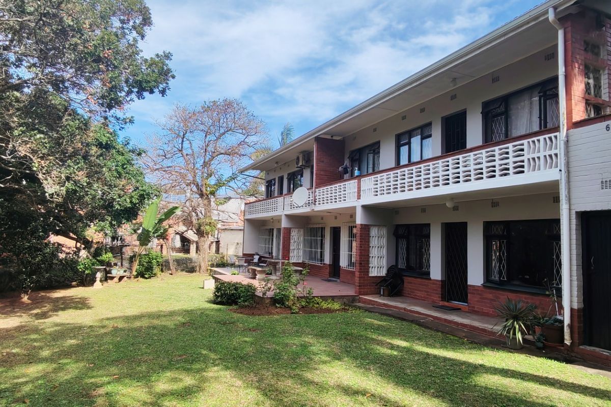 Rear view of a multi-story apartment building featuring a shared garden, ground floor patio, and upper-level balcony with security features.