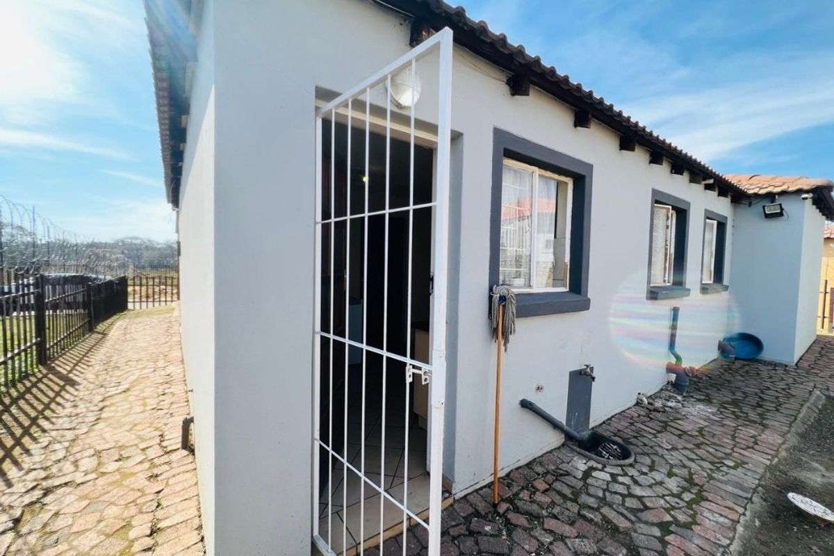 Rear exterior view of the property featuring a security gate, paved walkway, and exposed external plumbing along the wall.
