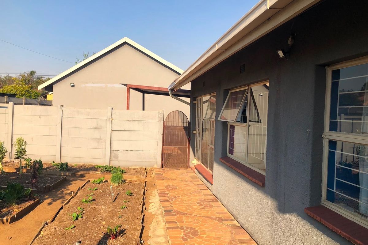 Rear exterior view of the property featuring a paved walkway, garden beds, and a secure boundary wall with a metal gate.