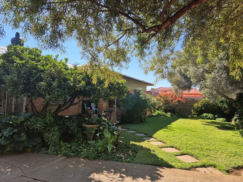 A well-maintained garden featuring a lush lawn, mature trees, stone stepping stones, and a view of the brick house exterior.
