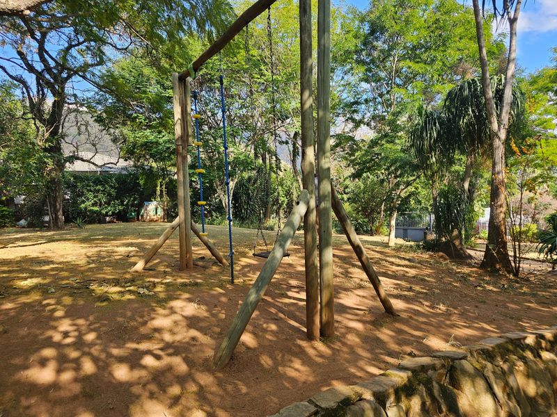 A spacious garden area featuring mature trees, a timber swing set, and a stone retaining wall.