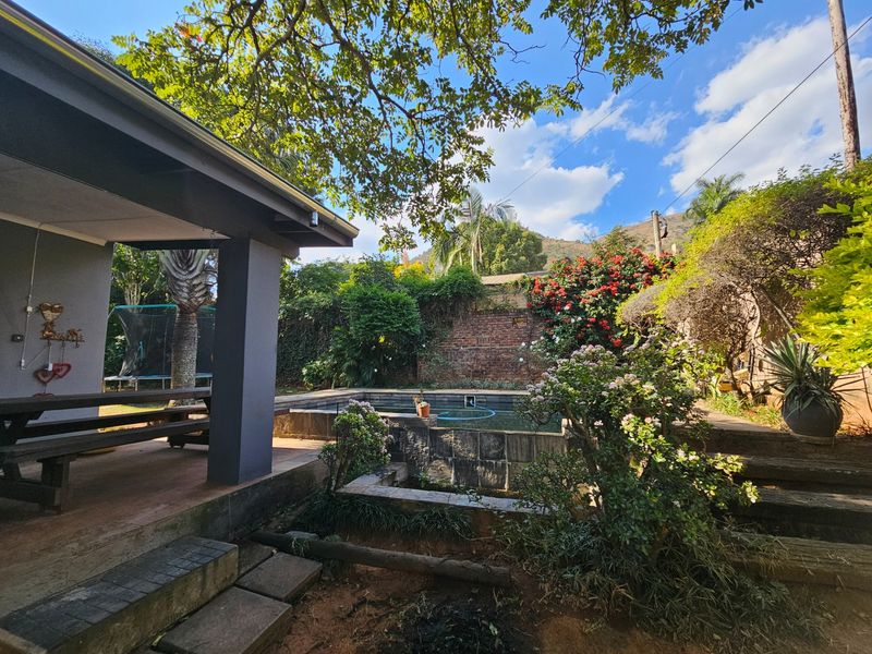 Outdoor pool area featuring a covered patio, stone retaining walls, and established garden landscaping with a brick boundary wall.
