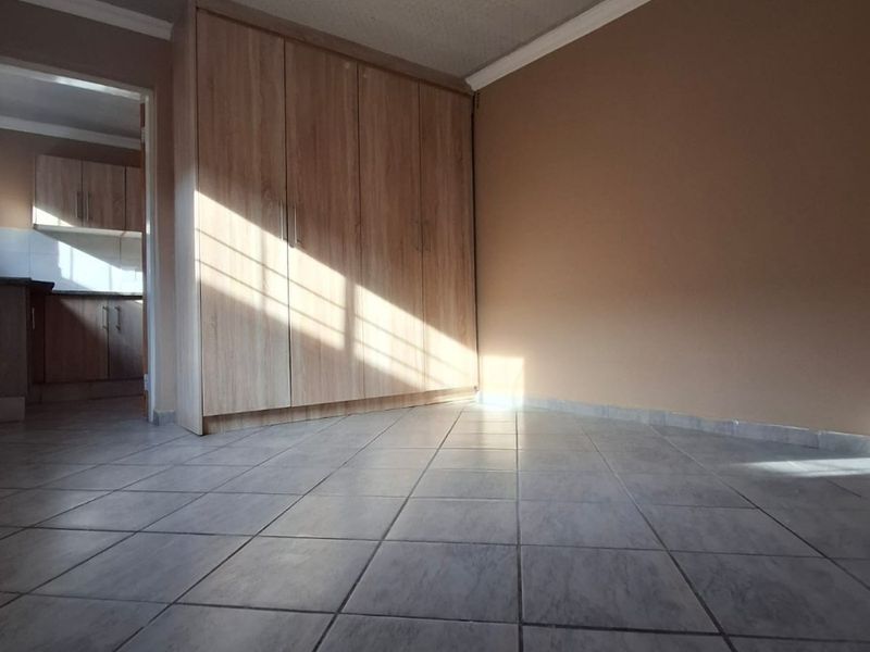 A bright, empty bedroom featuring tiled flooring, a built-in wardrobe, and coved ceilings, with a view into the adjacent kitchen area.