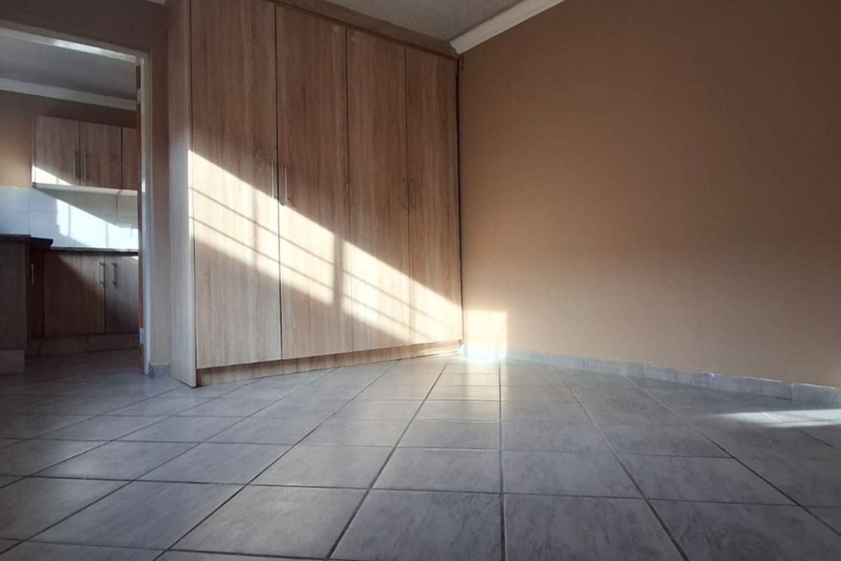 A bright, empty bedroom featuring tiled flooring, a built-in wardrobe, and coved ceilings, with a view into the adjacent kitchen area.