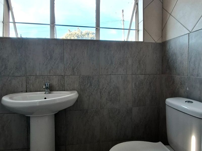 A functional bathroom featuring a pedestal basin, standard toilet, and neutral ceramic wall and floor tiling with a top-hung window for natural light.