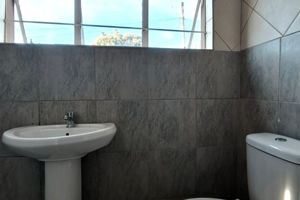 A functional bathroom featuring a pedestal basin, standard toilet, and neutral ceramic wall and floor tiling with a top-hung window for natural light.