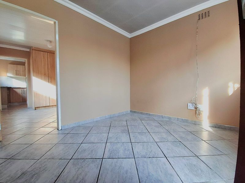 An empty living area featuring tiled flooring, neutral wall paint, and decorative cornice, with a view into the adjacent kitchen area.