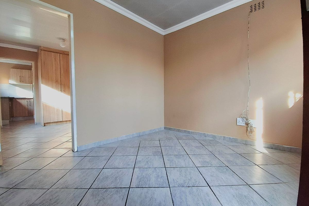 An empty living area featuring tiled flooring, neutral wall paint, and decorative cornice, with a view into the adjacent kitchen area.