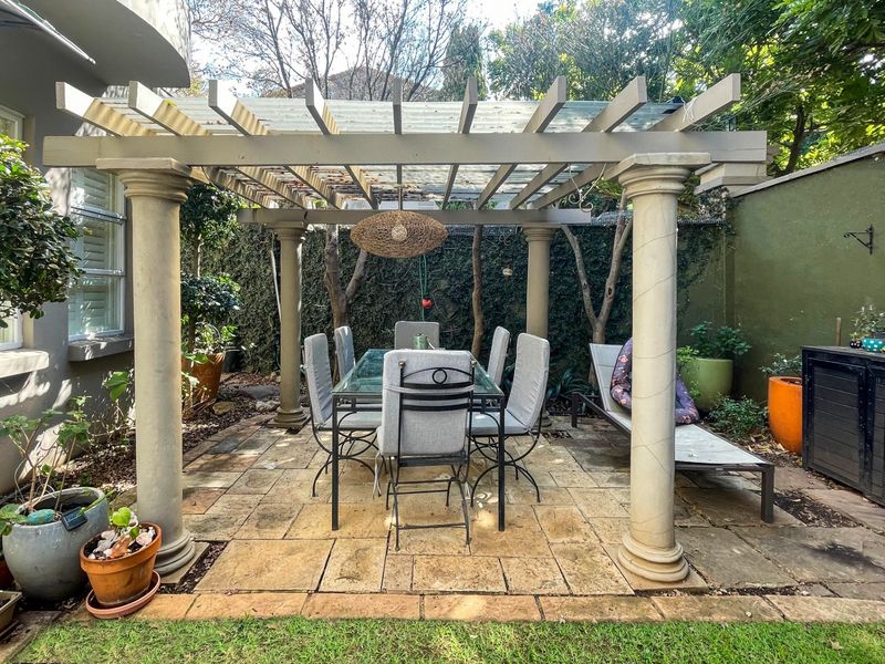 A covered outdoor patio area featuring a pergola structure with columns and stone paving, providing a shaded space for outdoor dining.