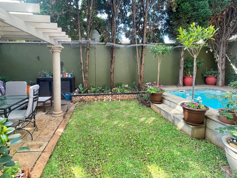 A private outdoor pool area featuring a paved patio with a pergola, a small lawn, and a raised garden bed against a boundary wall.