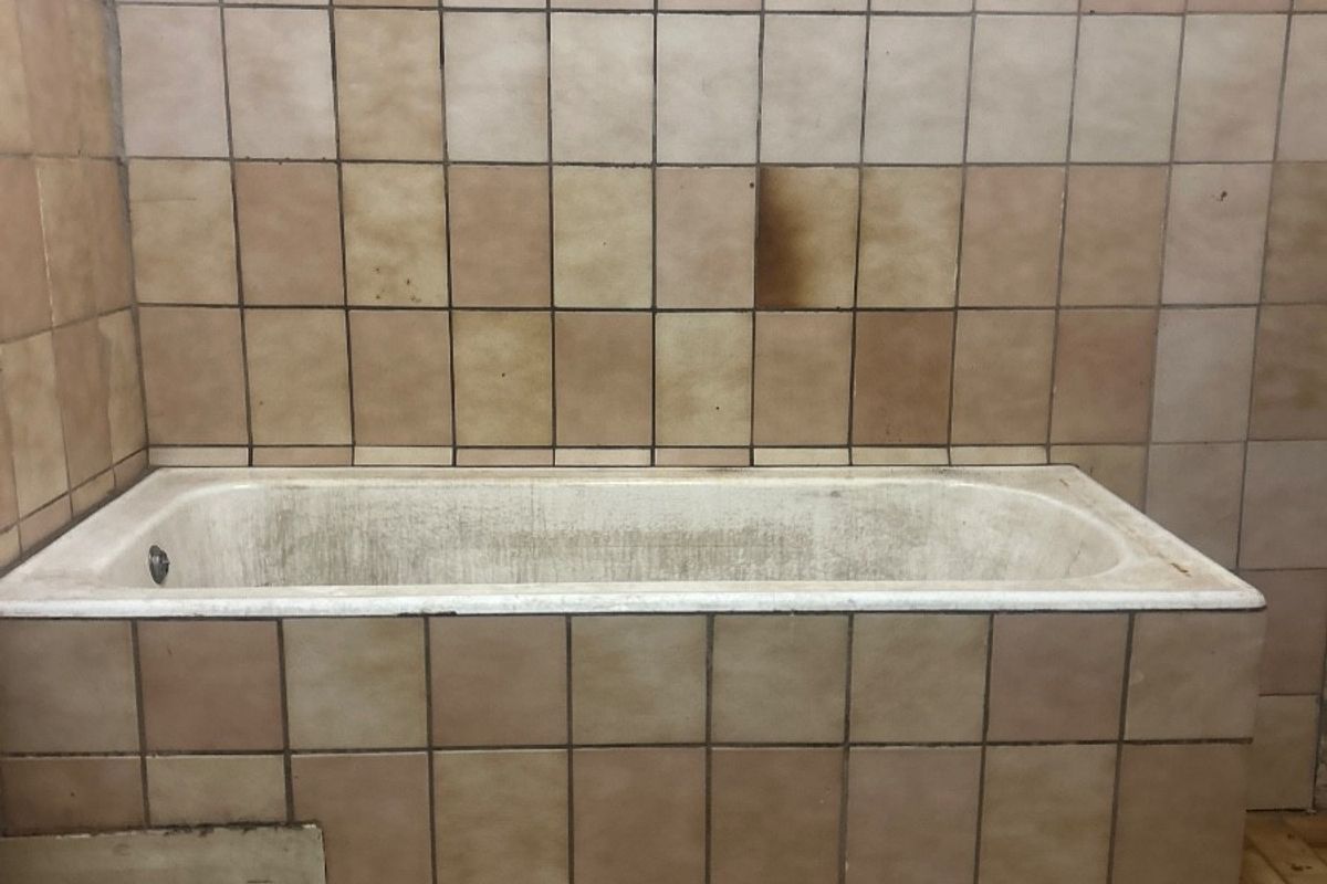 A dated bathroom featuring a built-in bathtub with ceramic tiling, showing significant signs of wear, staining, and surface damage requiring renovation.