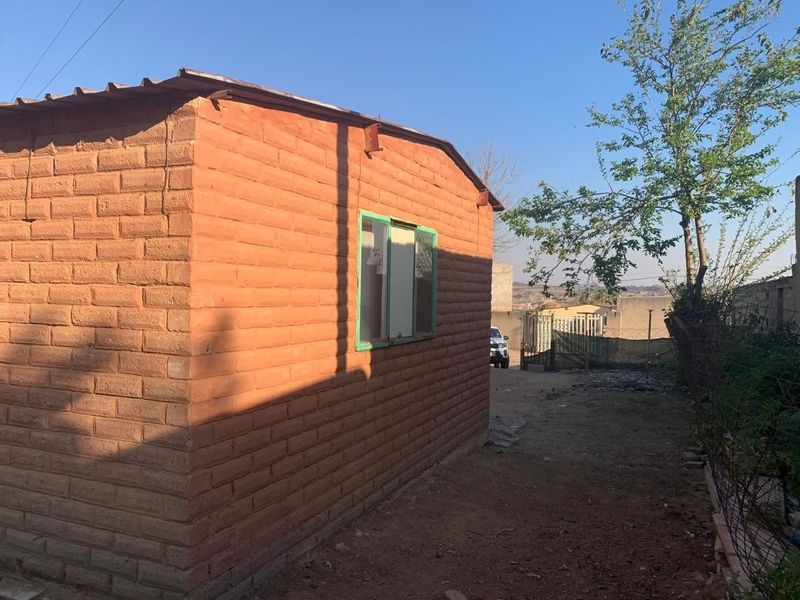 Rear view of a simple brick structure with a metal roof and sliding window, situated on an unpaved plot.