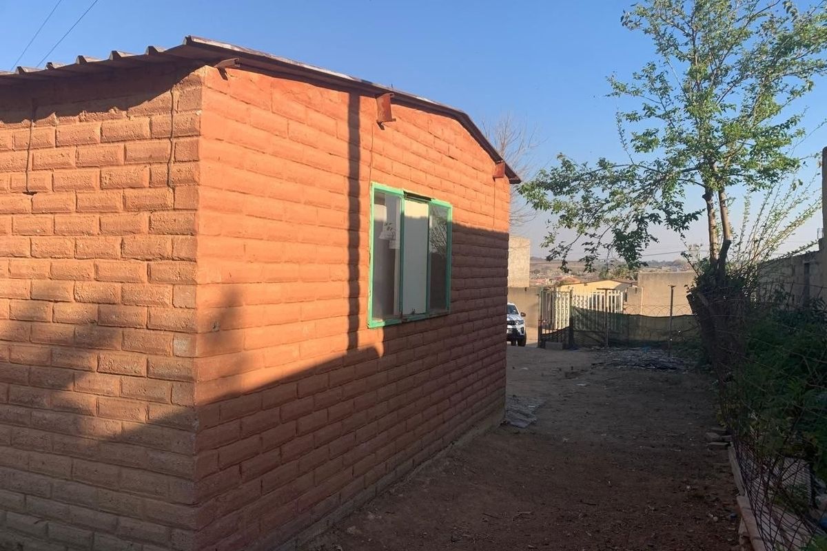 Rear view of a simple brick structure with a metal roof and sliding window, situated on an unpaved plot.