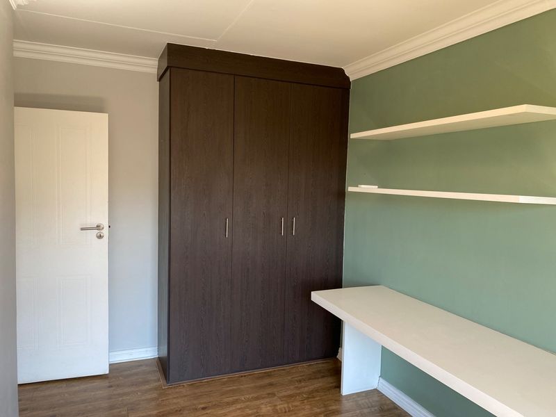A clean bedroom featuring a dark wood built-in wardrobe, a built-in desk, and floating shelves against a green accent wall.