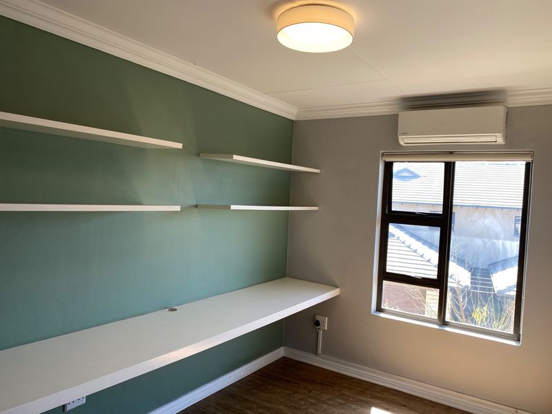 A bright home office featuring a built-in desk, floating shelving, laminate flooring, and a wall-mounted air conditioning unit.