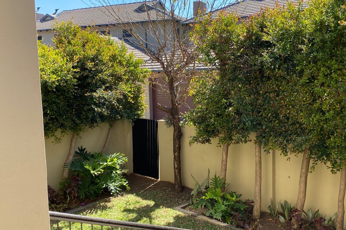 A private rear garden area featuring a well-maintained lawn, established trees, boundary walls, and a secure metal pedestrian gate.