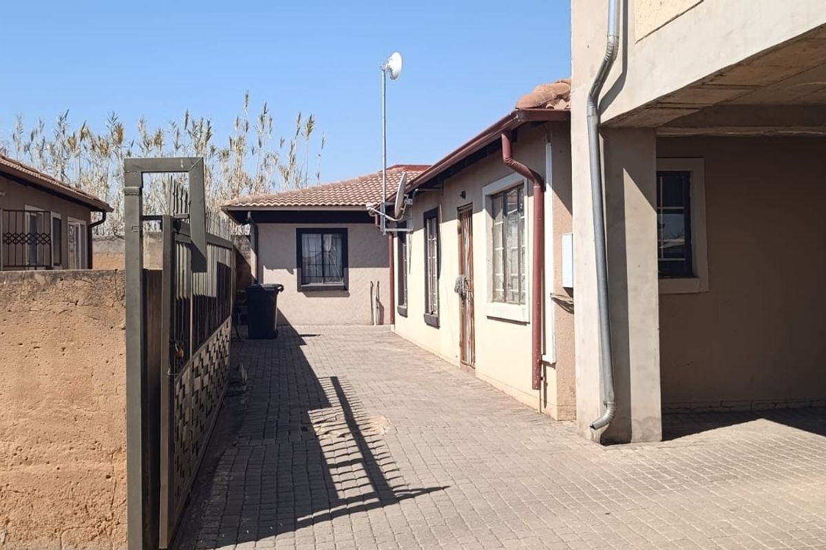 A view of the paved rear exterior area featuring a metal security gate, tiled roofing, and boundary walling.