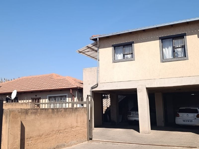 Front view of a double-storey house featuring a paved driveway, covered parking area, and a secure boundary wall with a metal gate.
