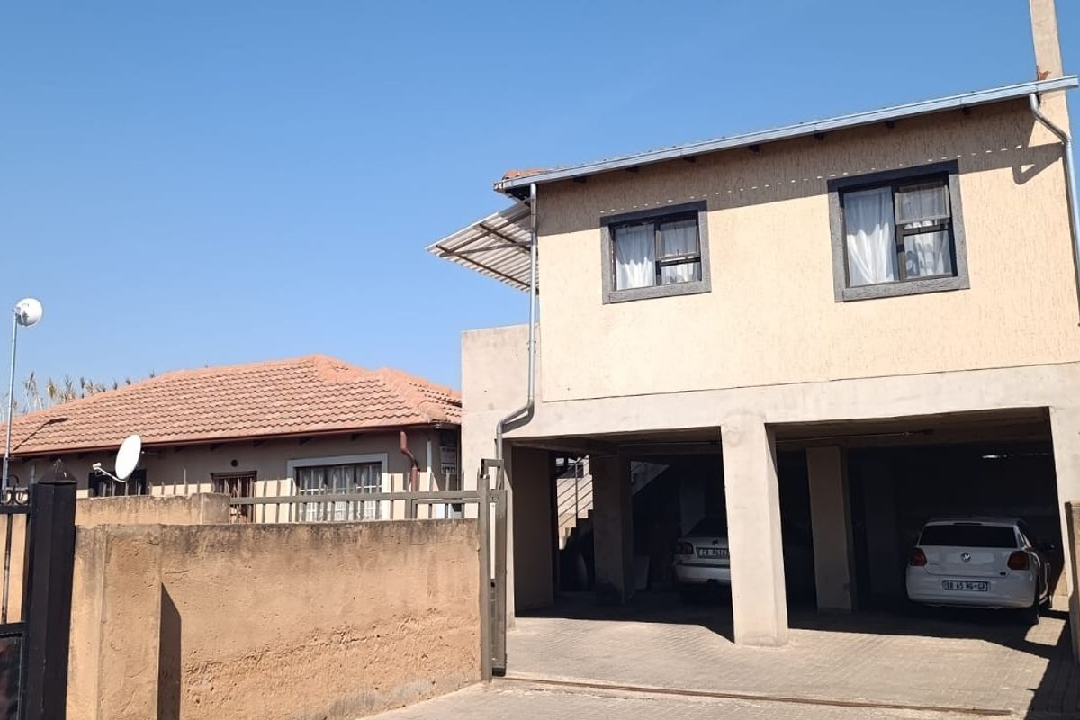 Front view of a double-storey house featuring a paved driveway, covered parking area, and a secure boundary wall with a metal gate.